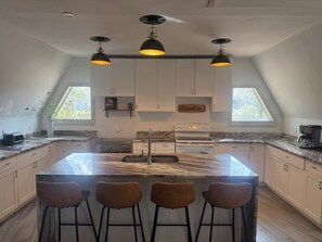 A modern kitchen with a large island, bar stools, pendant lighting, and a skylight. - Deland Dome Home- 98" TV - Open Kitchen (DeLand)