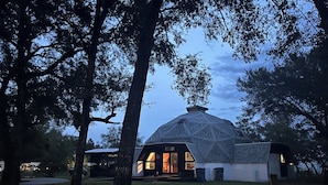 A large geodesic dome structure with a central entrance and surrounding trees. - Deland Dome Home- 98" TV - Open Kitchen (DeLand)