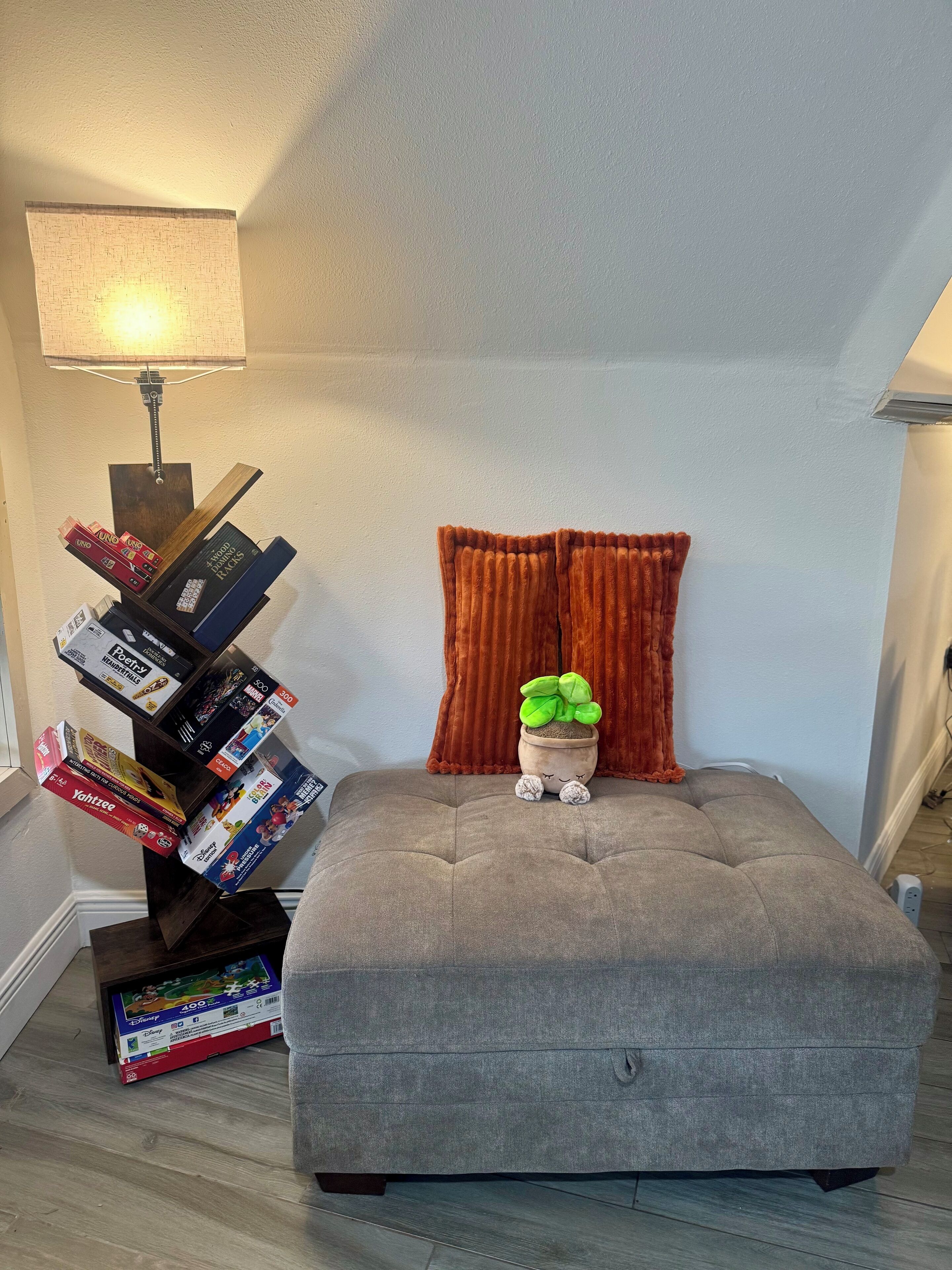 A cozy room with a grey tufted ottoman, a small potted plant, and a stack of board games.