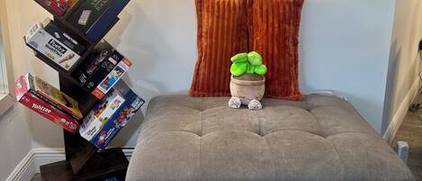 A cozy room with a grey tufted ottoman, a small potted plant, and a stack of board games.
