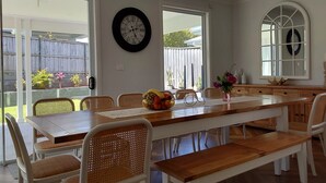 Un espace repas avec une longue table en bois, des chaises blanches et un banc. Deux horloges sont accrochées aux murs, un luminaire est fixé au plafond, et de grandes fenêtres offrent une vue sur le jardin.