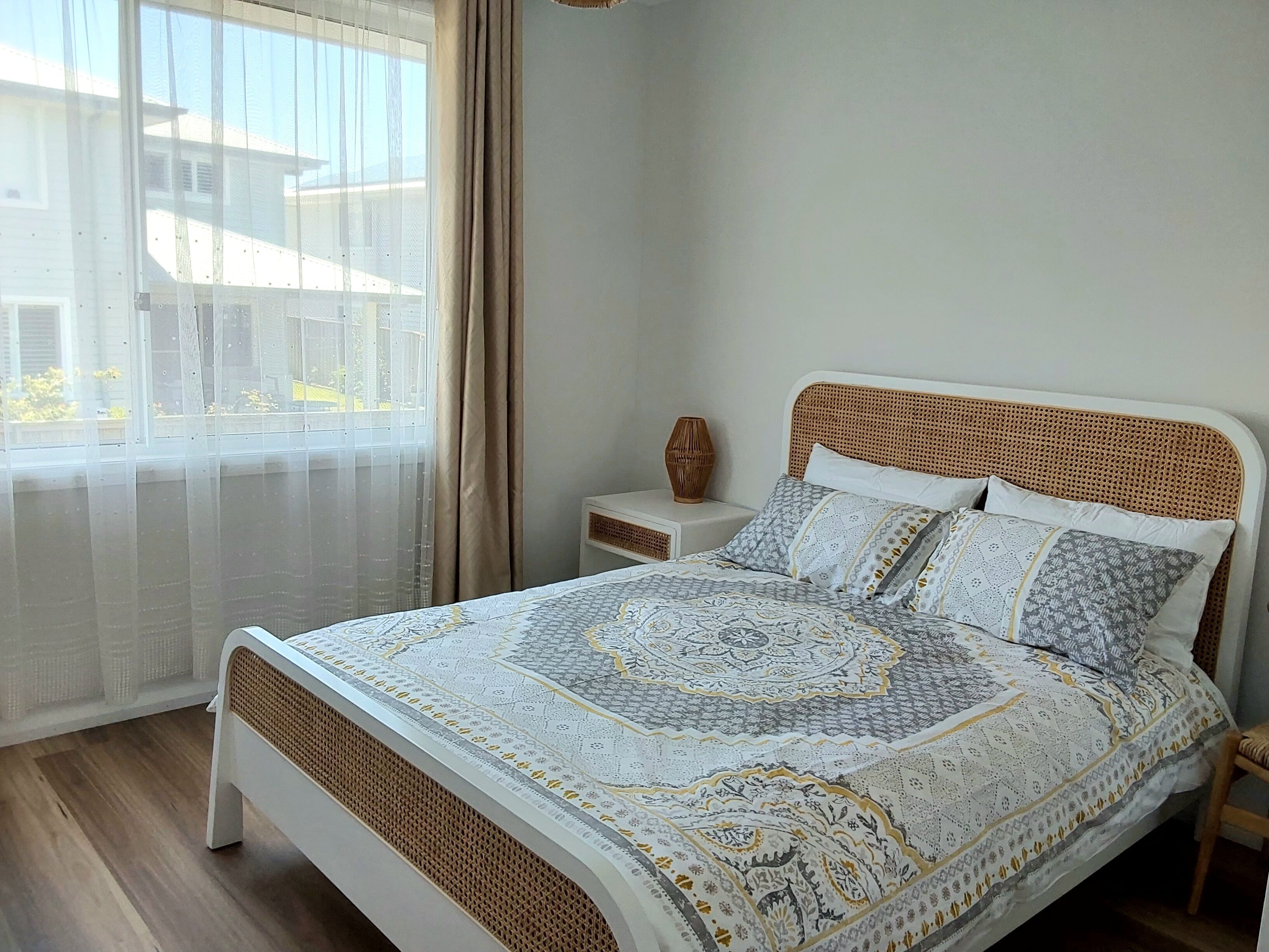 A bedroom with a bed, a nightstand, a window with sheer curtains, and a view of the outside.