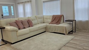 A spacious living room with a large L-shaped sofa, a small side table, and a large window with blinds. - Modern & open 3-bedroom house with 3 stall garage in a perfect location.. (Ventura)