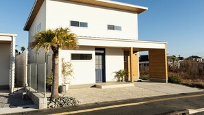 A modern two-story house with a flat roof, a covered porch, and a small garden with a palm tree. - Villa with a Natural Spring Bath and Wood-Fired Sauna — Aoshima (宮崎市青島)