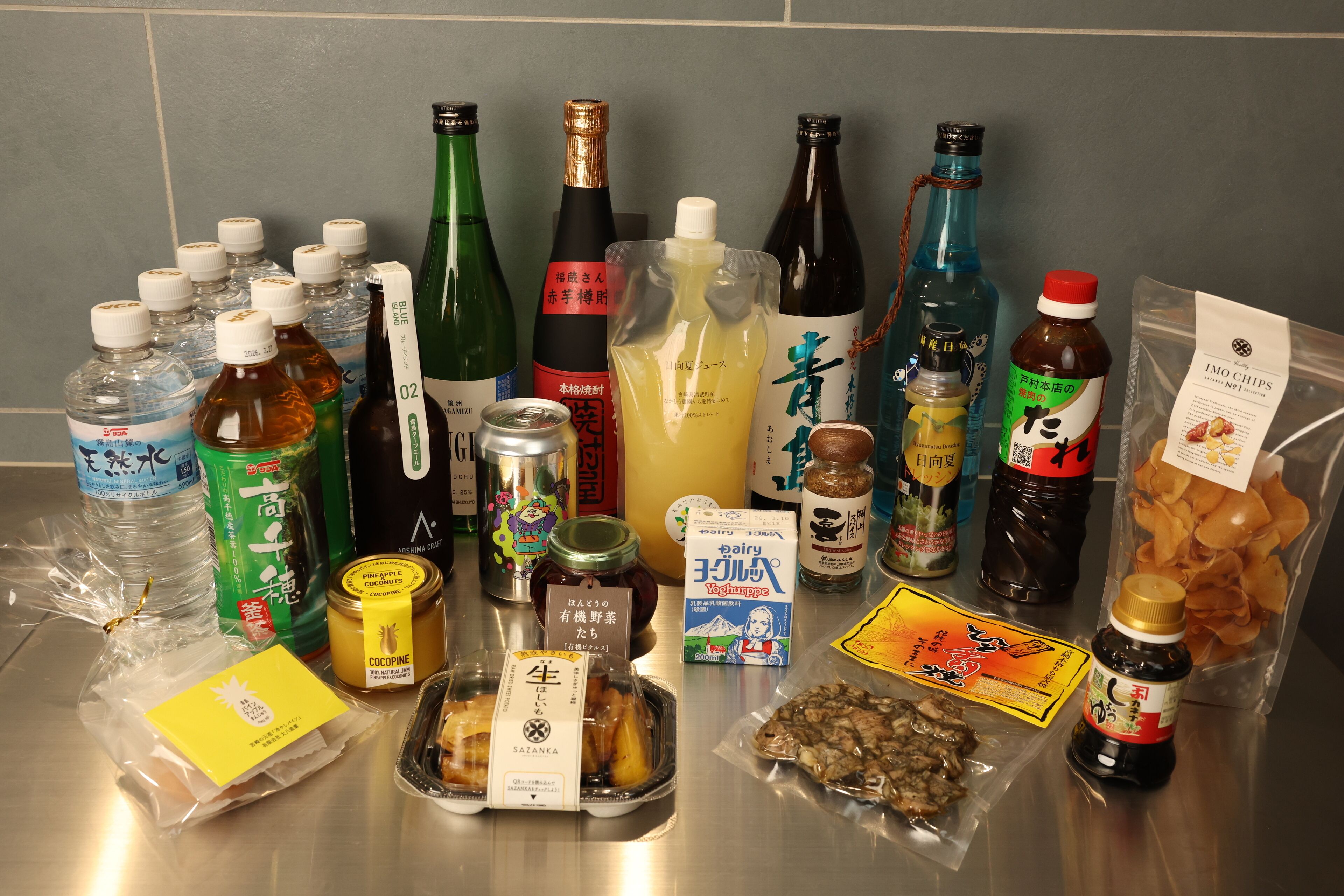 Diverse dranken en etenswaren, waaronder flessen, blikken en verpakte producten, op een metalen oppervlak gerangschikt.