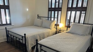A room with two beds, wooden flooring, and windows with blinds.