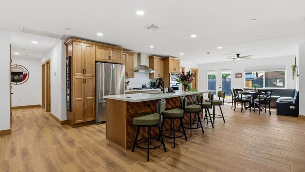 Eine moderne Küche mit Holzschränken, einem Edelstahlkühlschrank, einer zentralen Kochinsel mit Barhocker und einem Essbereich mit Tisch und Stühlen.