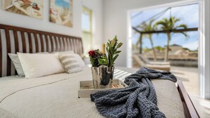 Ein Schlafzimmer mit einem Bett, einem Nachttisch mit Blumen und Blick auf Palmen und einen Strand durch das Fenster.