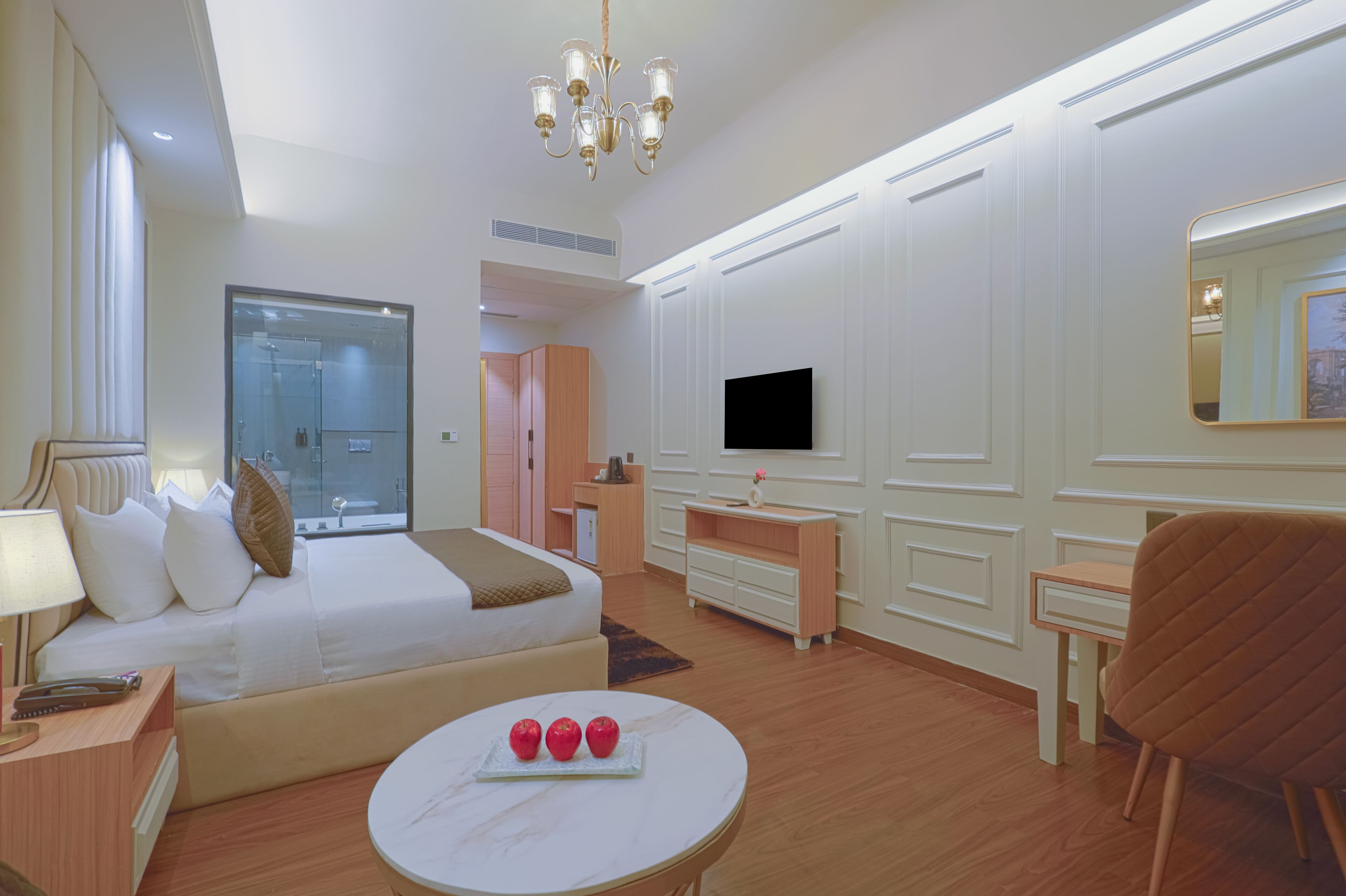 Een hotelkamer met een bed, nachtkastje, lamp, ronde tafel met appels, een televisie, spiegel en stoel.
