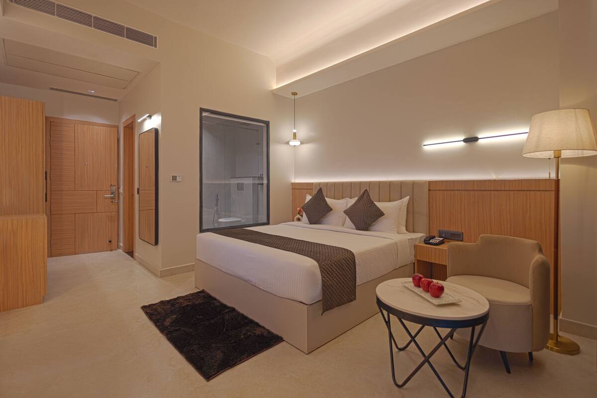 A modern hotel room with a large bed, a bedside table with a lamp, a small round table with apples, and a chair.
