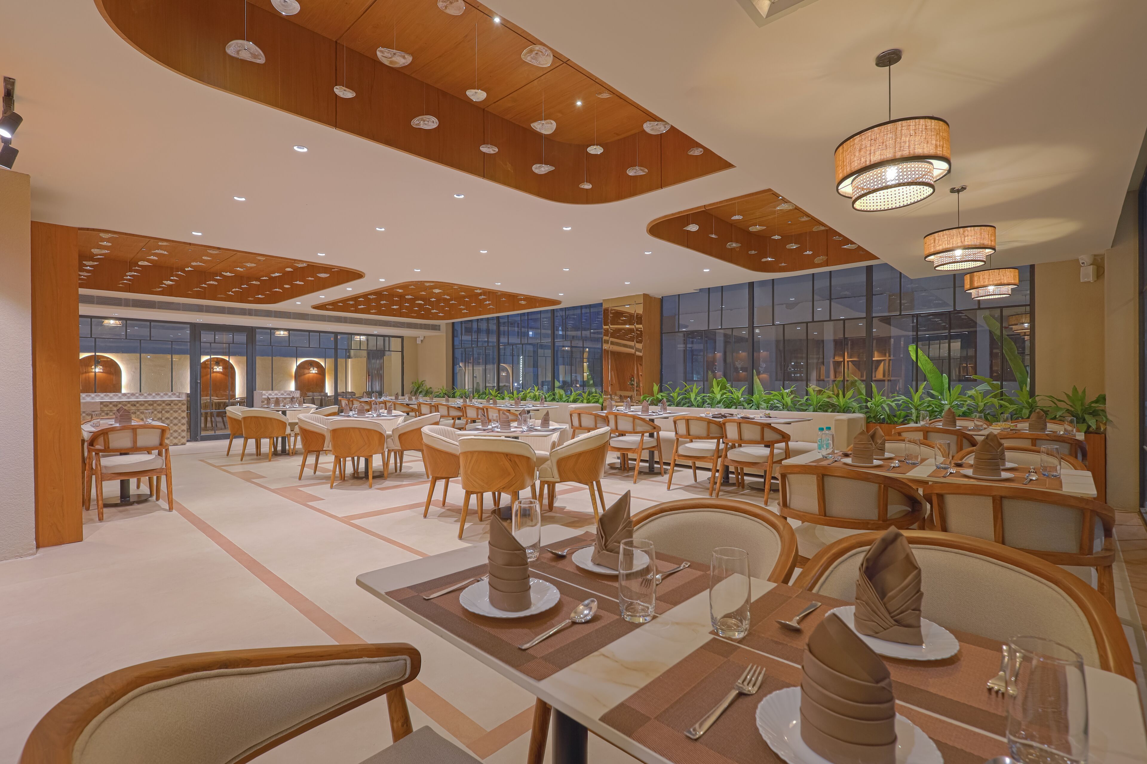 A spacious dining area with multiple tables and chairs, featuring a modern ceiling design and large windows with a view of greenery outside.
