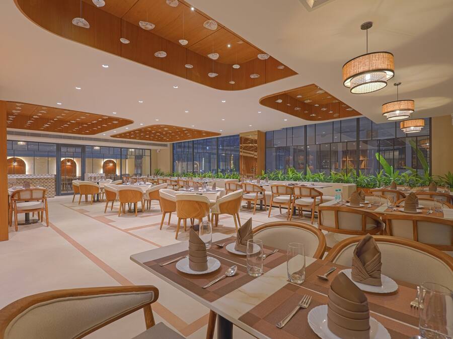 A spacious dining area with multiple tables and chairs, featuring a modern ceiling design and large windows with a view of greenery outside.