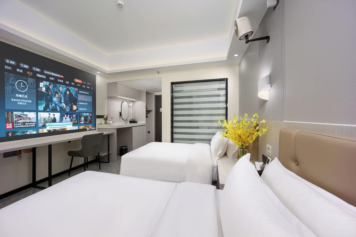 Habitació d'hotel amb dos llits, una taula de treball, un televisor que mostra un menú i un ram de flors grogues.