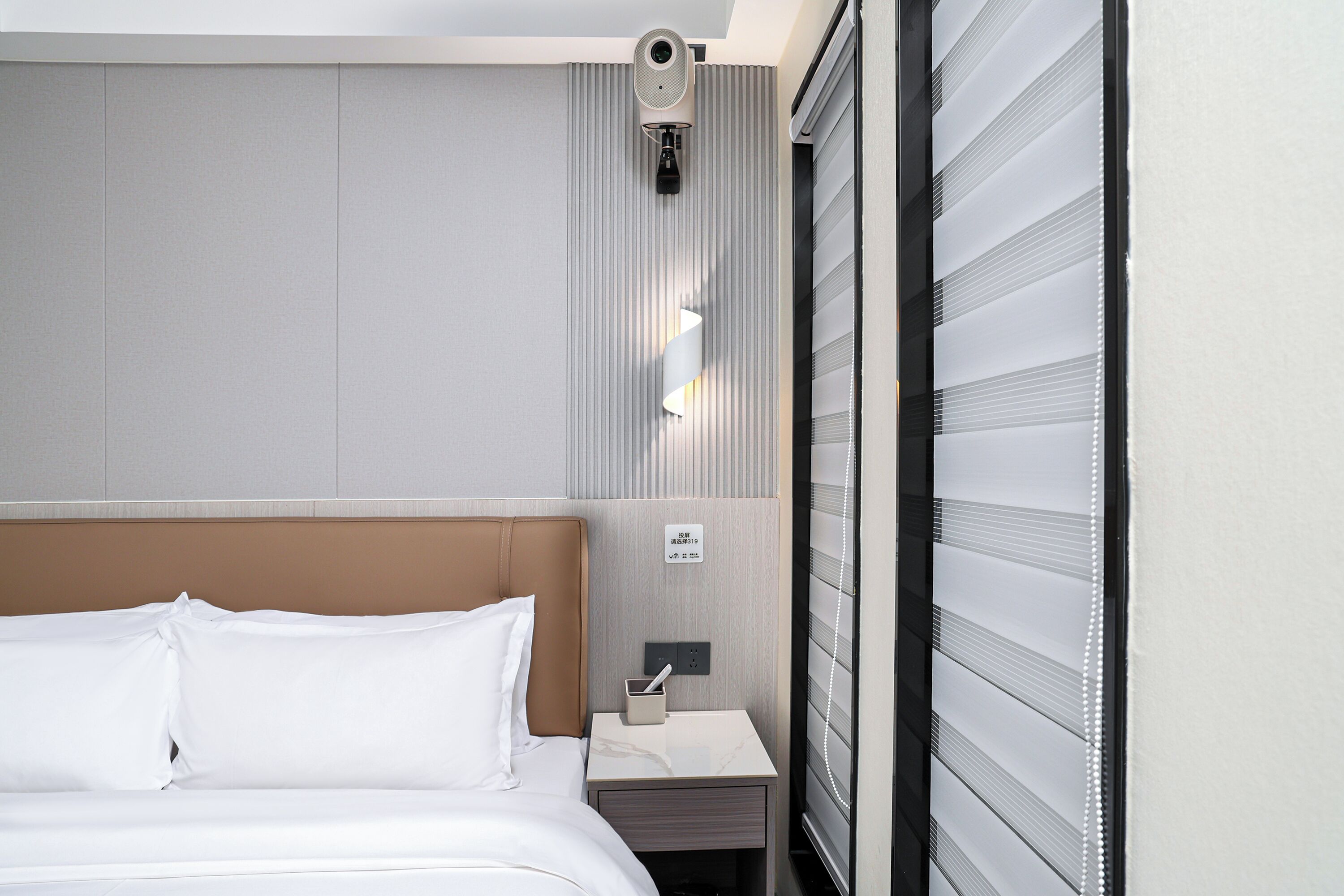 Habitación de hotel con cama, mesita de noche, cámara de seguridad en la pared y ventana con persianas.