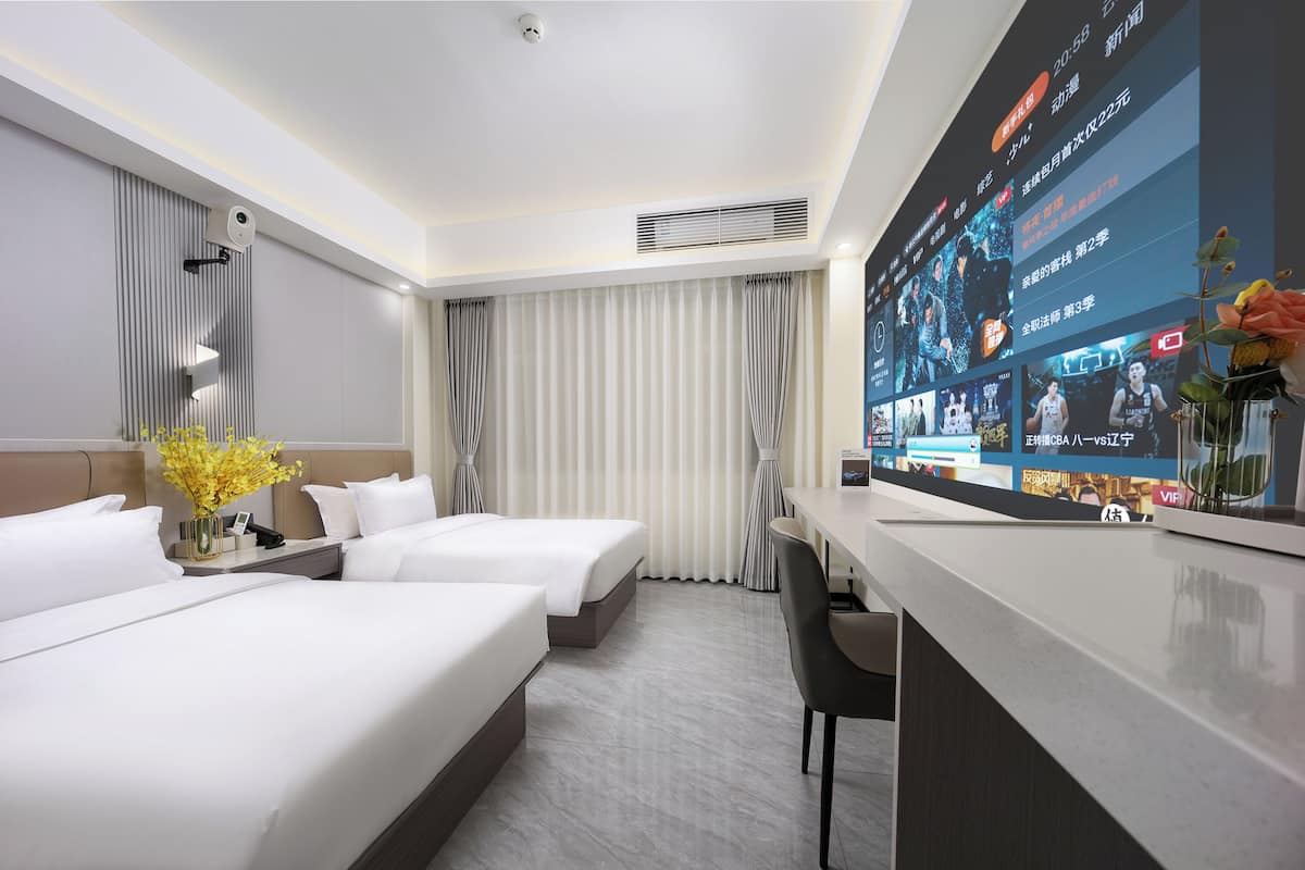 Habitació d'hotel amb dos llits, una taula de treball, una gran pantalla digital que mostra contingut, i un ram de flors.