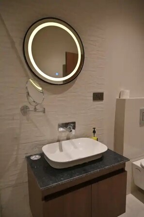 A bathroom with a round mirror, a white sink, and a toilet. - Hotel Phantom (Ganganagar)