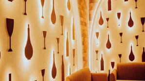 A cozy interior with a brick wall and a patterned wall design featuring wine glasses. There are cushioned seating areas and a table. - Hotel Phantom (Ganganagar)