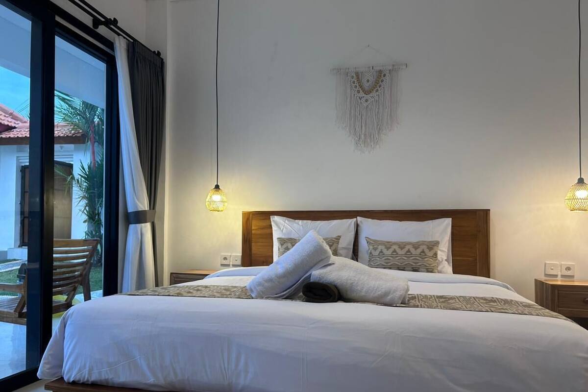 Een net opgemaakt bed met witte lakens en opgerolde handdoeken, een houten hoofdbord en twee bedlampen.
