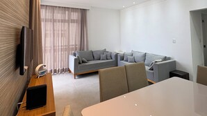 A modern hotel room with a large white table, two sofas, a TV mounted on the wall, and a large window with curtains.