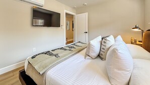 Ein Schlafzimmer mit Bett, Flachbildfernseher an der Wand, Nachttischlampe und einer Tür zu einem anderen Raum.