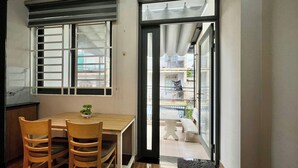 A room with a wooden dining table and chairs, a small potted plant, and a view of a balcony with a bench and a white chair. - AP Home (Ho Chi Minh City)