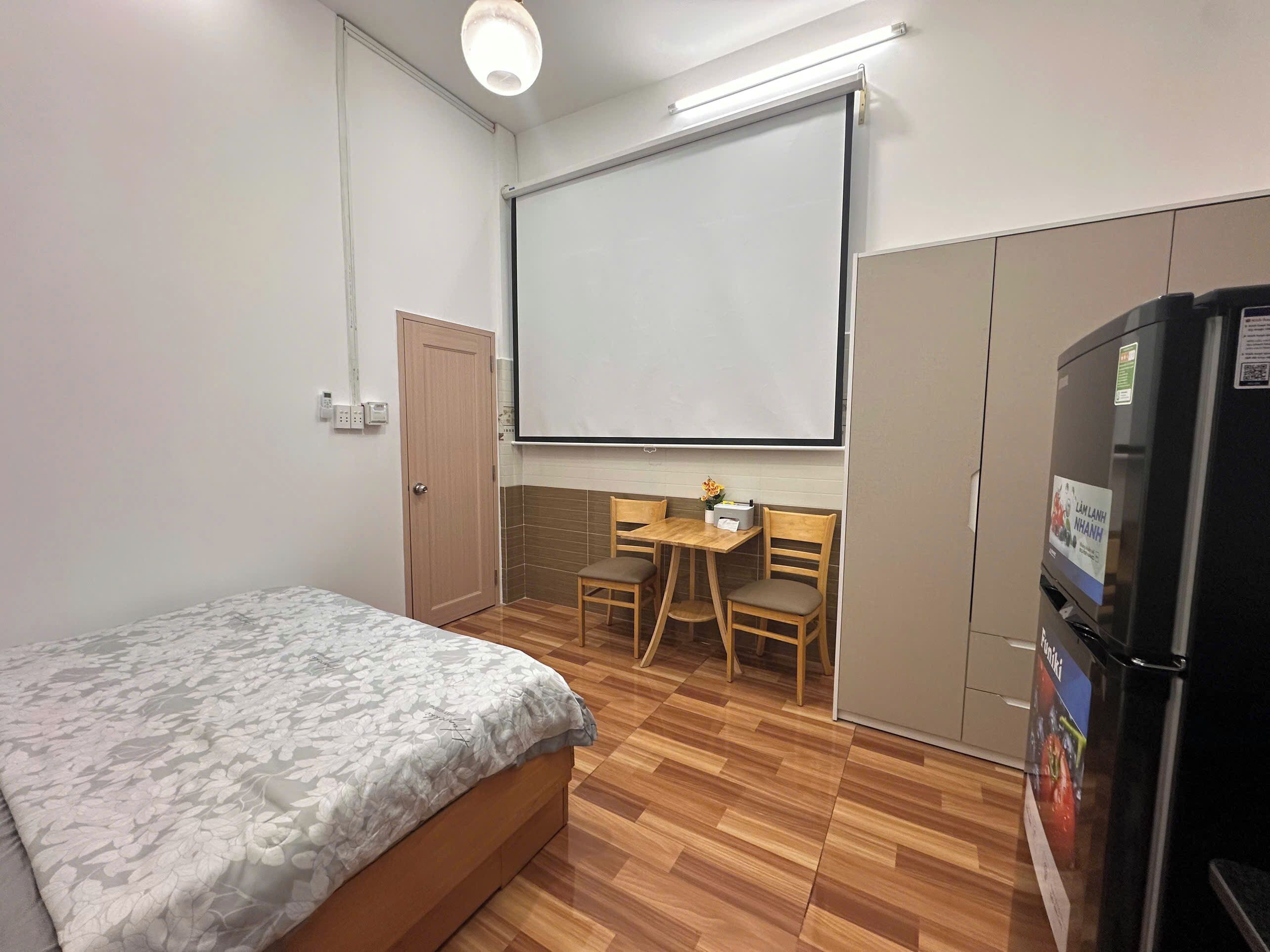 A room with a bed, a desk, a chair, a projector screen, a refrigerator, and a door.