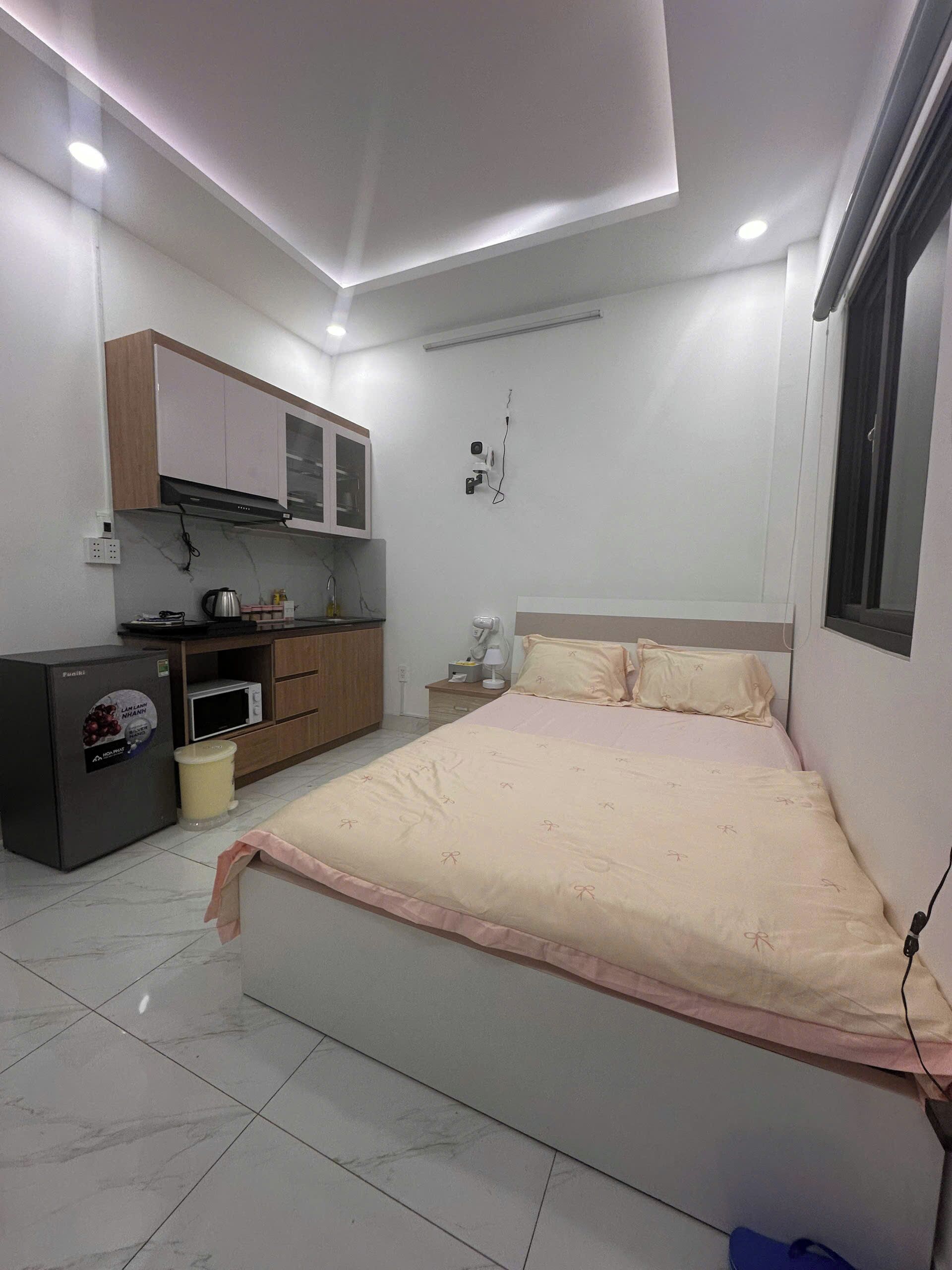 A bedroom with a bed, a bedside table, a nightstand, a wardrobe, a built-in microwave, and a refrigerator.