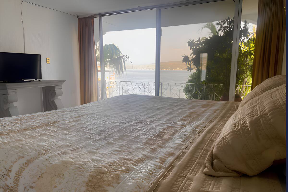 A bedroom with a large bed, a television on a stand, and a view of the sea through a sliding door.