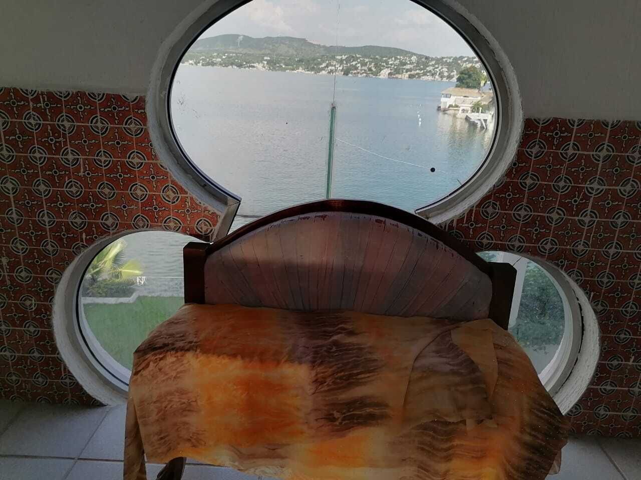 A room with a bed facing a large window with a view of a body of water and distant land.