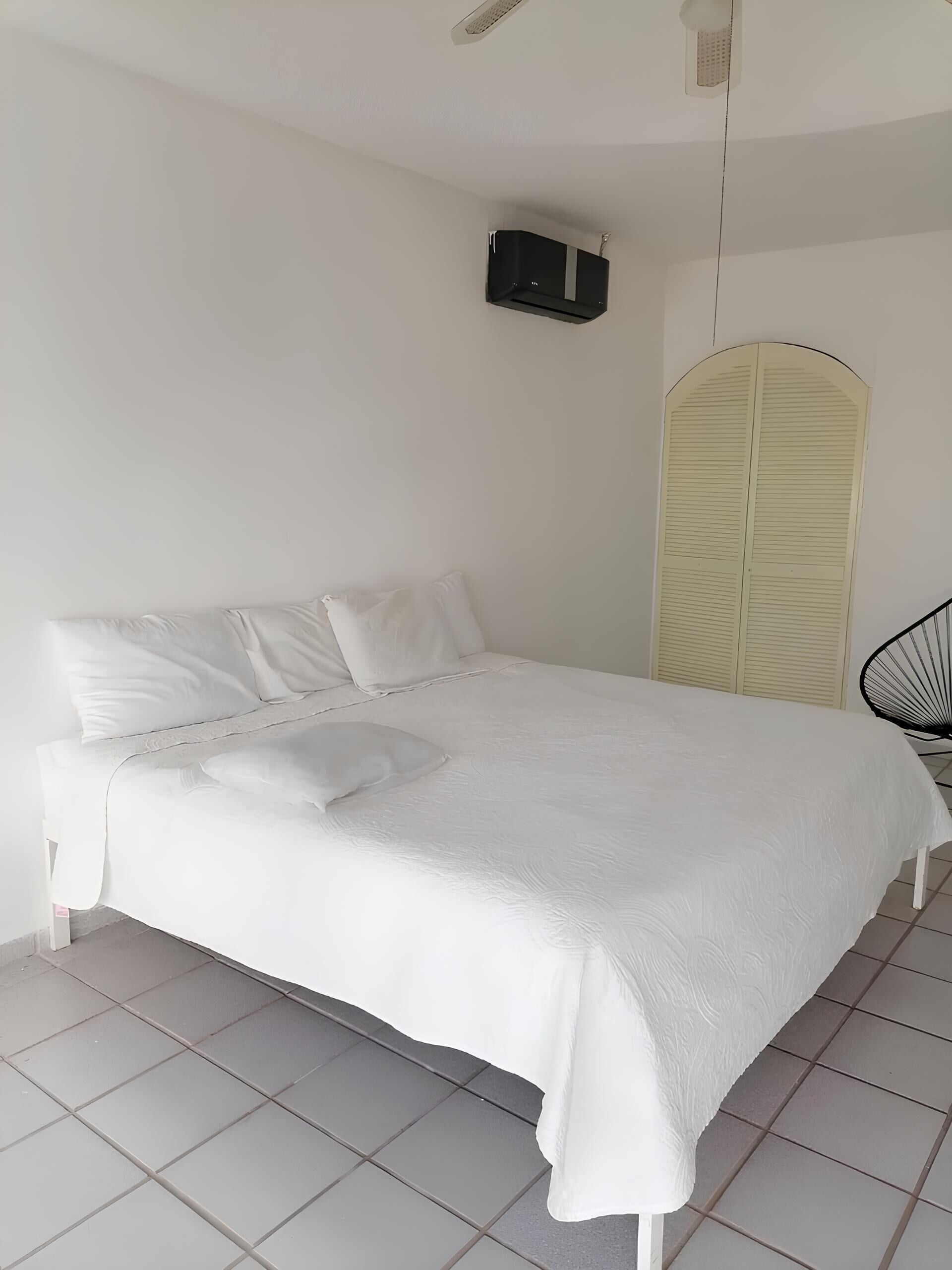 Een net opgemaakt bed met witte beddengoed, een zwart wandapparaat en een witte boogvormige deur.