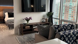 A modern living room with a large flat-screen TV mounted on the wall, a comfortable sofa with patterned cushions, a small side table with decorative items, and a view of tall buildings through large windows. - Amazing Views, Close to Football Stadium, Cruiseliners (Vancouver)