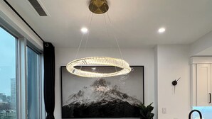 A modern dining area with a round table, four chairs, a chandelier, a painting, and a view of the city through large windows. - Amazing Views, Close to Football Stadium, Cruiseliners (Vancouver)