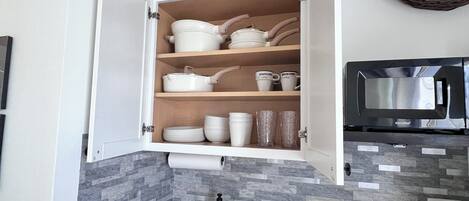 A kitchen with a black countertop, a stainless steel sink, a microwave, and a cabinet with dishes and cups.