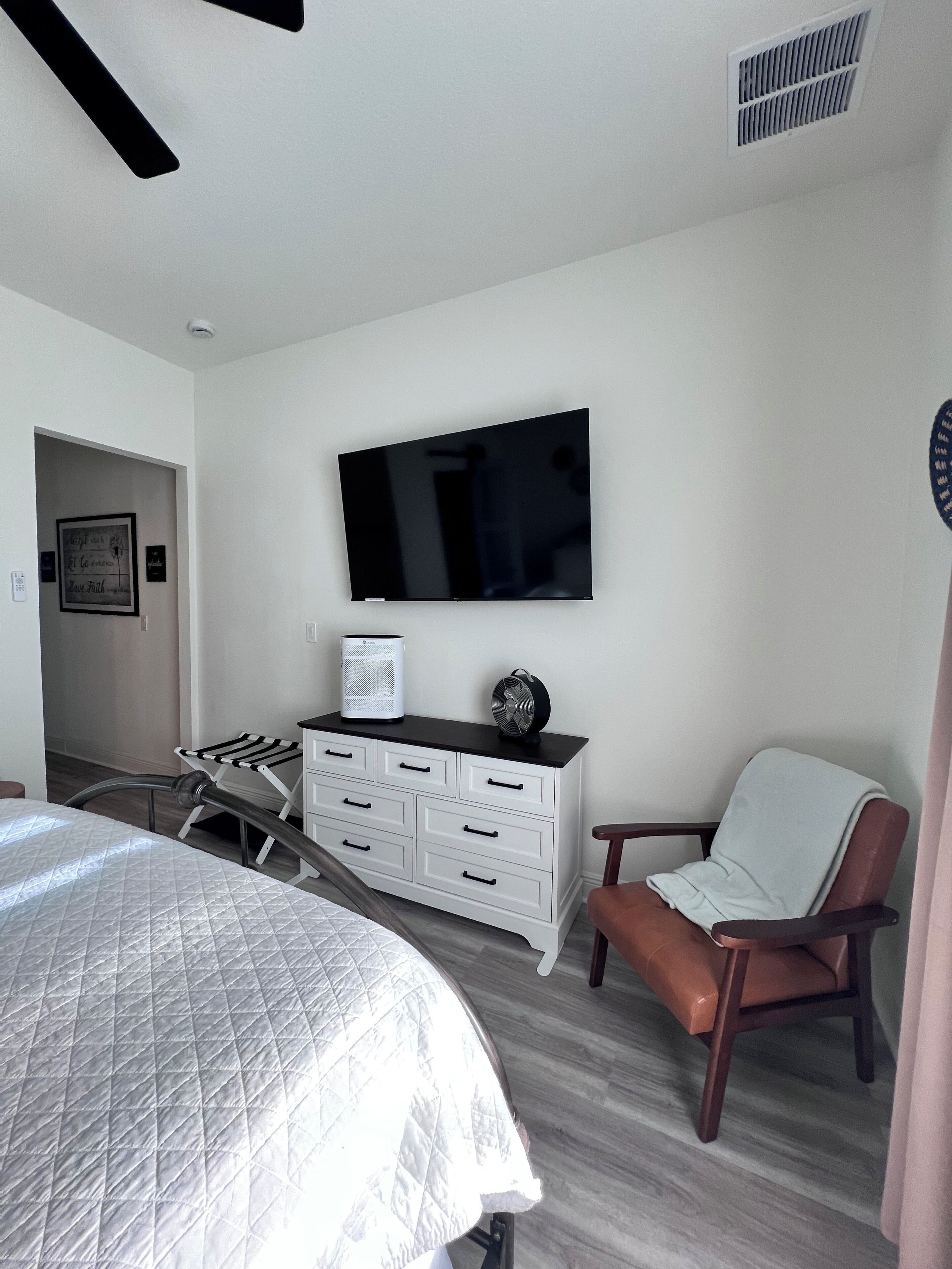 Une chambre à coucher comprenant un lit, une commode, une chaise, un téléviseur et une bouche d’aération.