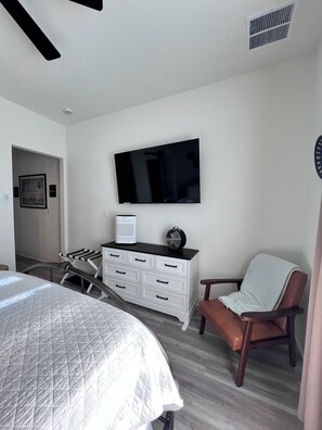 Une chambre à coucher comprenant un lit, une commode, une chaise, un téléviseur et une bouche d’aération.