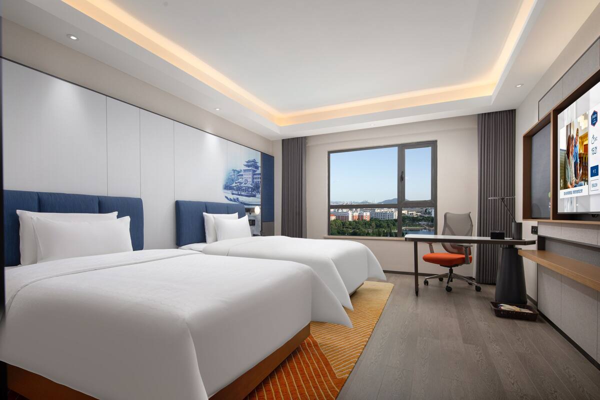 A hotel room with two beds, a desk, a chair, a television, and a large window with a city view.