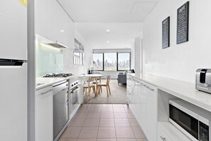 Eine moderne Küche mit weißen Schränken, einem Gasherd, einer Mikrowelle und einem Toaster. Es gibt einen Essbereich mit Tisch und Stühlen sowie ein großes Fenster mit Blick auf die Stadt.