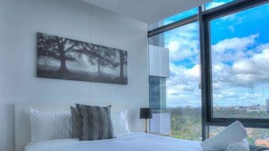 Ein modernes Schlafzimmer mit einem großen Bett, Blick auf die Stadt durch ein Fenster und einem Gemälde an der Wand.