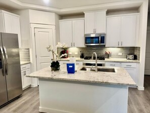 A modern kitchen with white cabinetry, a stainless steel refrigerator, a microwave, and a granite countertop. - FW002 | Ft. Worth Retreat | 3BD |Garage |Workspace (Fort Worth)