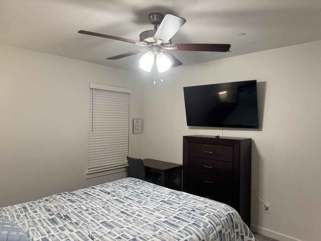 A bedroom with a bed, a desk, a chair, a dresser, a ceiling fan, and a flat-screen TV.