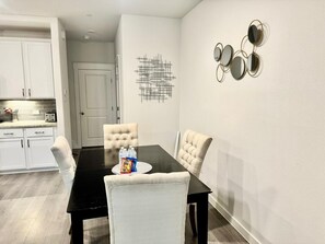 Uma área de refeições com uma mesa preta, cadeiras brancas estofadas e uma porta branca ao fundo.