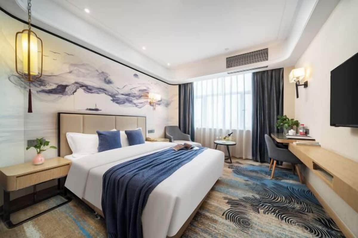 Ein Hotelzimmer mit Bett, Nachttisch, Lampe, Schreibtisch, Stuhl und einem großen Fenster mit Vorhängen.