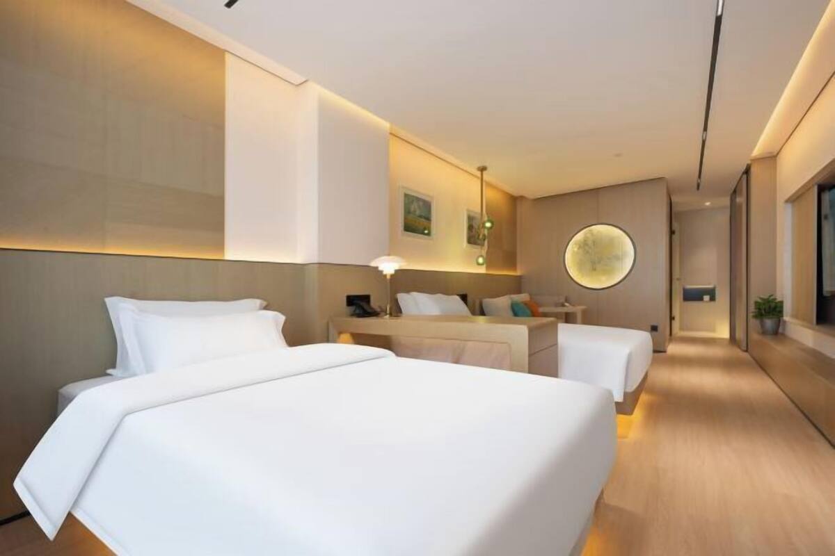 Ein Hotelzimmer mit zwei Betten, einem Nachttisch, einer Lampe, einer wandmontierten runden Leuchte und einer Pflanze.