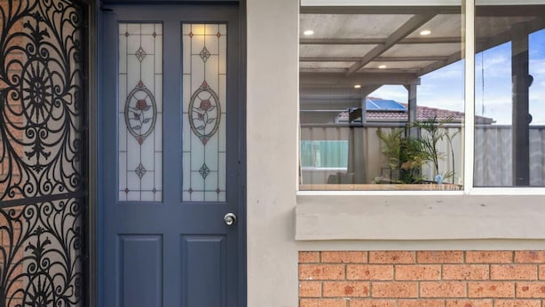 A blue door with a glass panel and a decorative metal gate, a brick wall, and a covered outdoor area with a window.