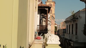 Een smalle straat met gebouwen aan weerszijden, een balkon met een witte balustrade, en een heldere blauwe lucht.