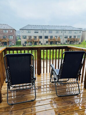 Une terrasse en bois avec deux chaises noires et une rambarde, donnant sur un immeuble d’habitation doté de nombreux balcons.