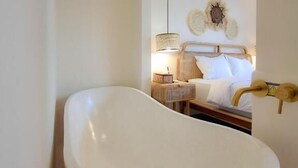 Een moderne badkamer met een vrijstaand bad, een grote spiegel en een nachtkastje met een lamp.