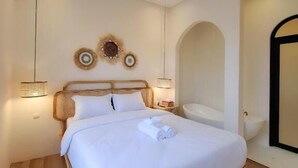 Een net opgemaakt bed in een hotelkamer met een houten hoofdbord, een wandairconditioner en een klein tafeltje met een lamp.