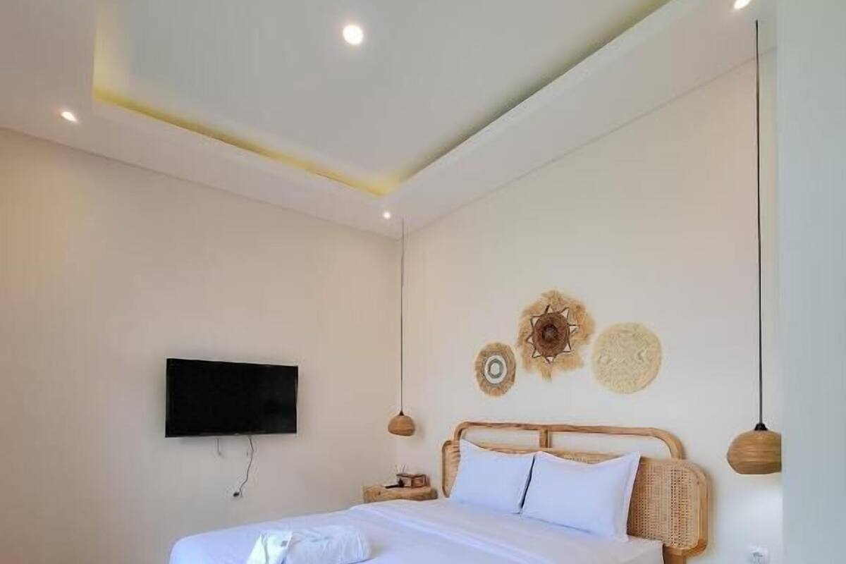 Un dormitorio con cama de madera, ropa de cama blanca, un televisor de pantalla plana montado en la pared y una mesita de noche con lámpara.