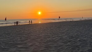 Cena de praia com o sol se pondo sobre o oceano, pessoas caminhando pela orla e bandeiras delimitando a área da praia.